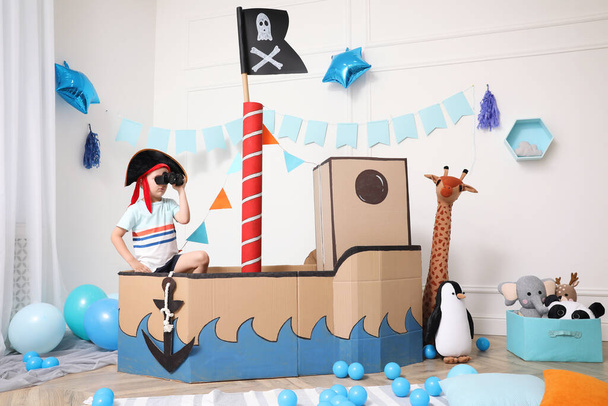 Little boy playing with binoculars in pirate cardboard ship at home. Child's room interior - Foto, imagen