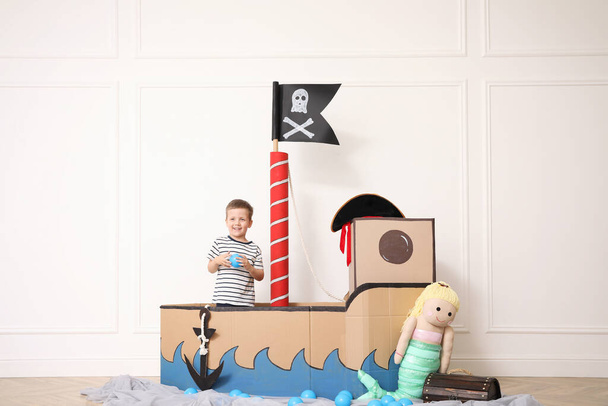 Cute little boy playing in pirate cardboard ship near white wall indoors - Foto, imagen
