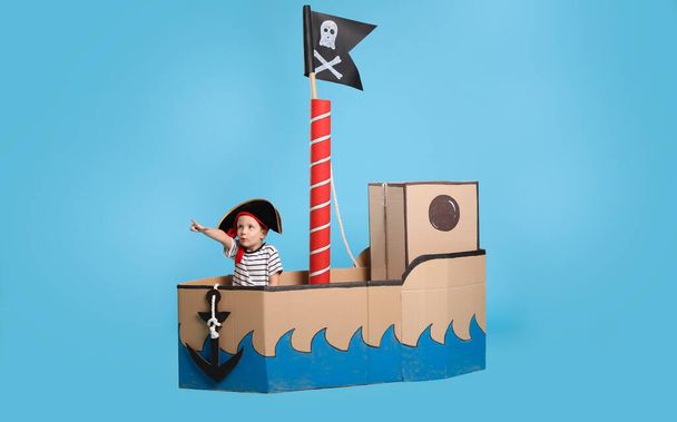 Cute little boy playing in pirate cardboard ship on turquoise background - Φωτογραφία, εικόνα