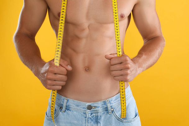 Shirtless man with slim body and measuring tape on yellow background, closeup - Φωτογραφία, εικόνα