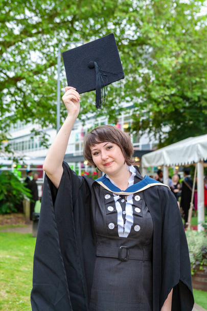 Estudiante con el pelo castaño usando un vestido de graduación de la universidad sonríe y sostiene su gorra sobre la cabeza. Oxford, Inglaterra - Foto, Imagen
