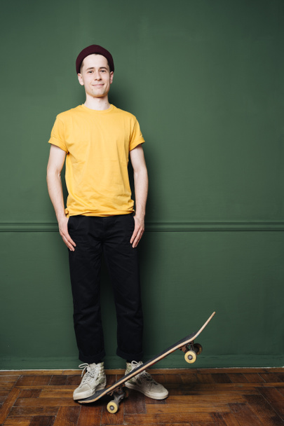 Homme caucasien en t-shirt jaune posant avec patin sur fond vert foncé mur - Photo, image