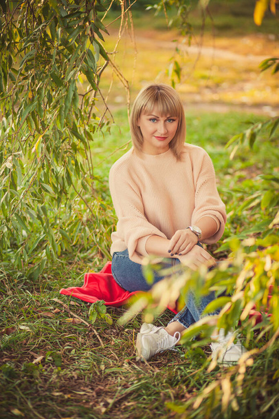 Portret blondynki z krótką fryzurą siedzącej na czerwonym kocu na polanie w lesie. Zewnętrzne spacery i odpoczynek w jesiennym lesie - Zdjęcie, obraz