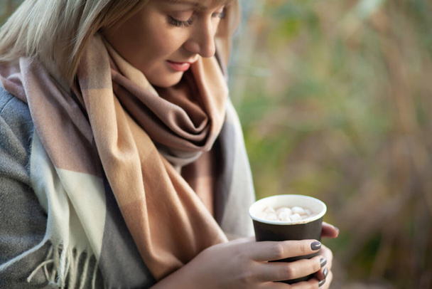 Blonde Frau mit Kurzhaarschnitt in grauem Wollmantel und kariertem Schal trinkt Kaffee-Cappuccino mit Marshmallows. Wandern und Picknick in der Natur im herbstlichen Wald - Foto, Bild