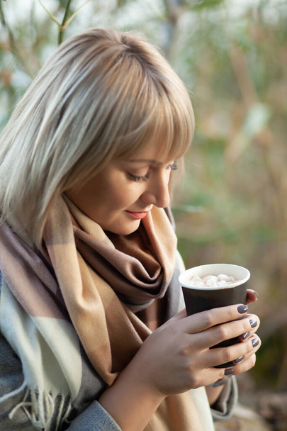 Блондинка с короткой стрижкой в сером шерстяном пальто и клетчатом шарфе пьет кофе капучино с зефиром. Прогулки и пикник на природе в осеннем лесу - Фото, изображение