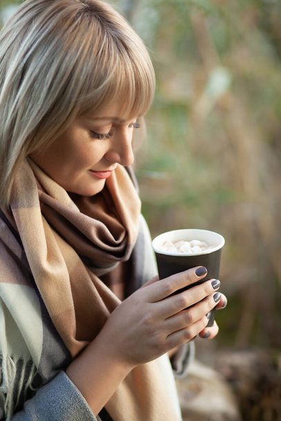 Femme blonde avec coupe de cheveux courte en manteau de laine grise et foulard à carreaux boit cappuccino café avec guimauves. Promenade et pique-nique dans la nature en forêt d'automne - Photo, image