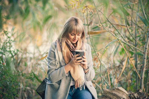 Blonde woman with short haircut in gray wool coat and plaid scarf drinks coffee cappuccino with marshmallows. Walking and picnic in nature in autumn forest - Photo, Image