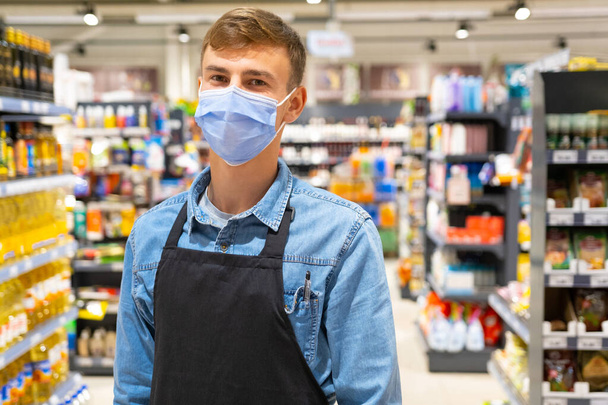 Loja trabalhador homem vestindo camisa jeans com manga comprida ter uma caneta no bolso do peito e avental cor preta posando em uma máscara facial em pé no meio do mercado da loja. Foto de alta qualidade - Foto, Imagem