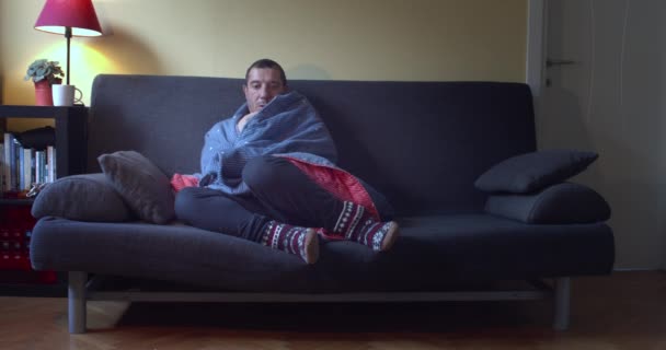 Hombre enfermo con fiebre sentado en el sofá envuelto en una manta azul utiliza aerosol nasal - Metraje, vídeo