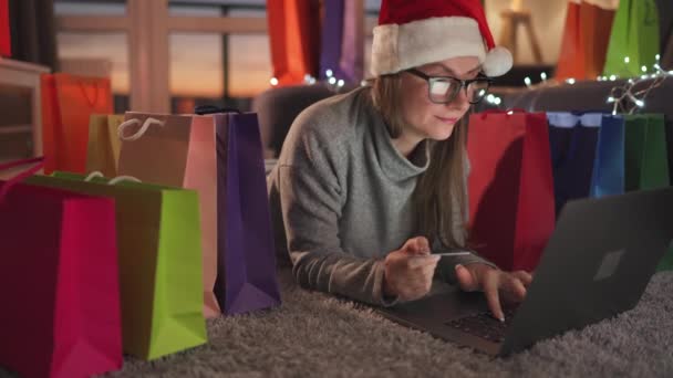 Mujer con gafas con un sombrero de Santa Claus está acostado en la alfombra y hace una compra en línea con una tarjeta de crédito y un ordenador portátil. Bolsas de compras alrededor. - Metraje, vídeo