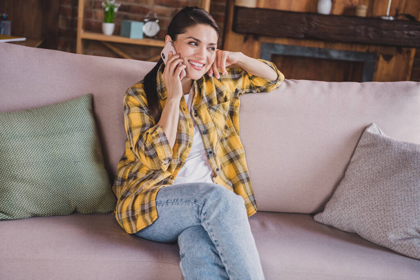 Photo of positive pretty person sit on couch speak telephone toothy smile have good mood enjoy weekend indoors - Фото, изображение