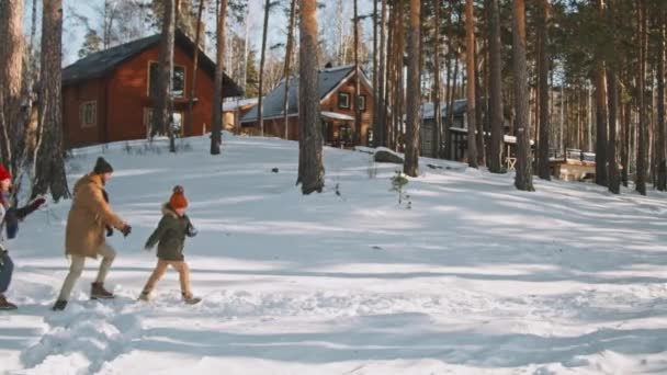 Güzel bir kış günü karın içinde koşan sıcak parkalar, şapkalar ve eşarplar giyen mutlu ebeveynler ve küçük kızlar. Arka planda ağaçlarla çevrili rahat kulübeler - Video, Çekim