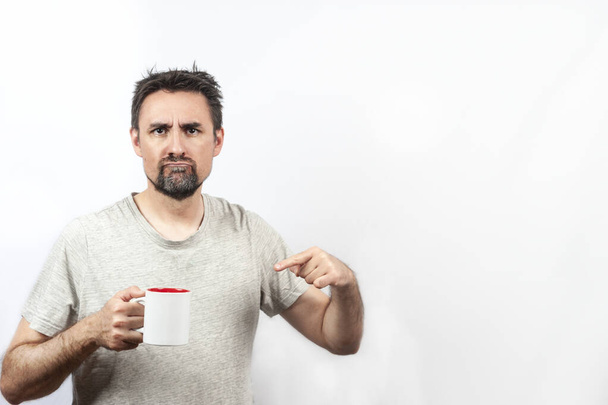 Dark haired, bearded man in gray shirt; with expression of wanting coffee or tea, with one hand holding a cup . Concept waking up. Copy space and white background - Photo, Image
