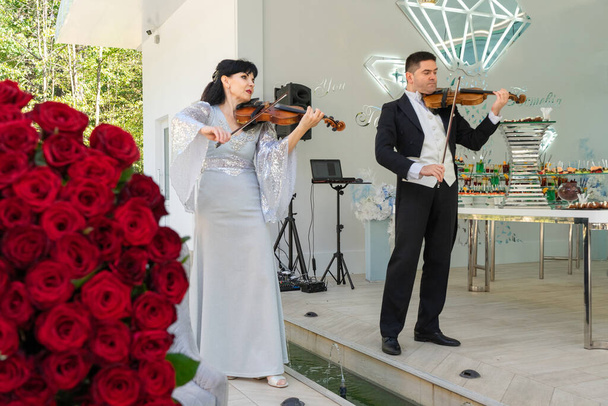 Houslista a houslista hrají na housle o aristokratickém svátku na pozadí velké kytice rudých růží. - Fotografie, Obrázek