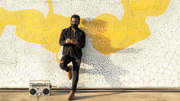 Jeune Afro homme utilisant un smartphone mobile tout en se tenant contre un mur urbain et en écoutant de la musique avec écouteurs et boombox vintage - Photo, image