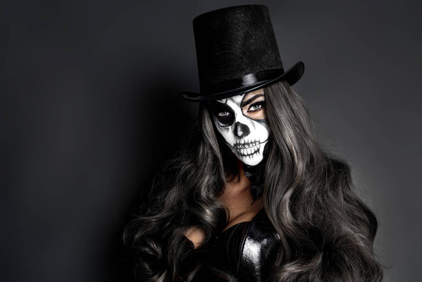 Mooi model in Halloween make-up op zwarte achtergrond. Sexy vrouw met hoge hoed en schedel make-up. Halloween make-up concept. - Foto, afbeelding
