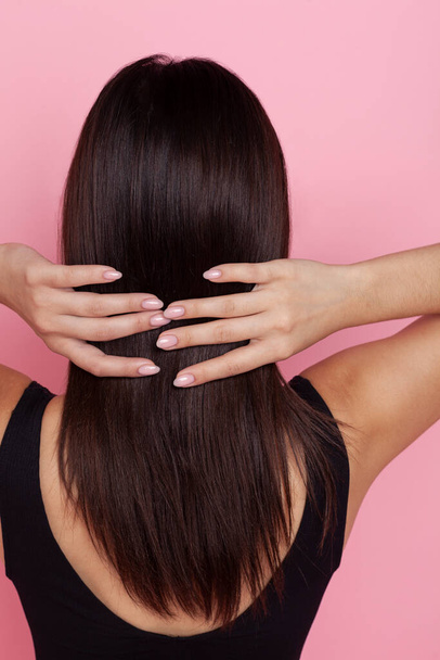 brunette with long well-groomed hair on a pink background stands with her back - Foto, Imagem