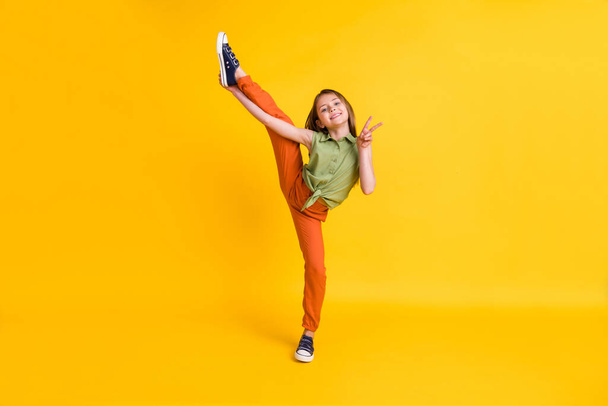 Longitud completa tamaño del cuerpo foto niña mostrando v-signo haciendo ejercicios deportivos aislado vibrante color amarillo fondo - Foto, Imagen