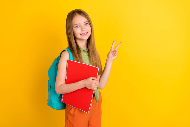 Photo portrait schoolgirl wearing green shirt rucksack keeping book stack showing v-sign isolated vivid yellow color background - Φωτογραφία, εικόνα