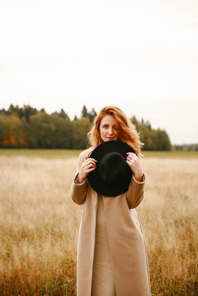Autumn portrait, a red-haired girl in a black hat and a brown coat walking in nature. Beautiful portrait in natural locations, blurred background and blurred focus - Foto, afbeelding