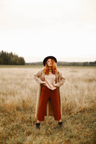 Autumn portrait, a red-haired girl in a black hat and a brown coat walking in nature. Beautiful portrait in natural locations, blurred background and blurred focus - Foto, imagen