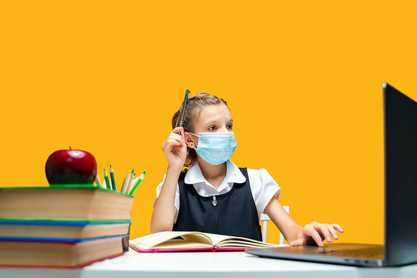  Schoolgirl raising hand and sitting at laptop wearing mask. Yellow background. Distance Learning - Фото, зображення