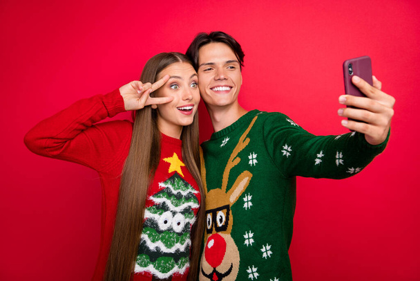 Foto van blij stel meisje show v-teken jongen nemen telefoon selfie dragen lelijk pullover geïsoleerde rode kleur achtergrond - Foto, afbeelding