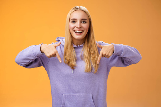 Amused joyful nice blond girlfriend pointing down present cool new product smiling broadly recommending try check out standing orange background happily grinning in hoodie - Fotoğraf, Görsel