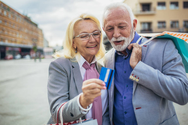 Happy mature couple walking with their shopping purchases on a sunny day using credit card. - Photo, Image