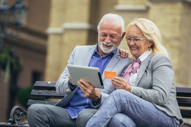 Couple âgé assis dans le parc sur banc et faire du shopping en ligne avec tablette et carte de crédit
 - Photo, image