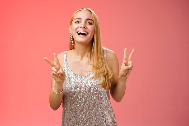 Funny carefree charming caucasian blond female model in silver stylish glamour dress show peace victory gesture smiling broadly have fun celebrating birthday party, standing red background - Photo, Image
