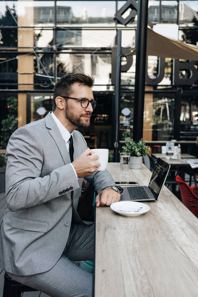 Middle age businessman sitting in cafe bar and enjoying at coffee break between two meetings. He uses laptop computer and he is positive and confident.  - 写真・画像