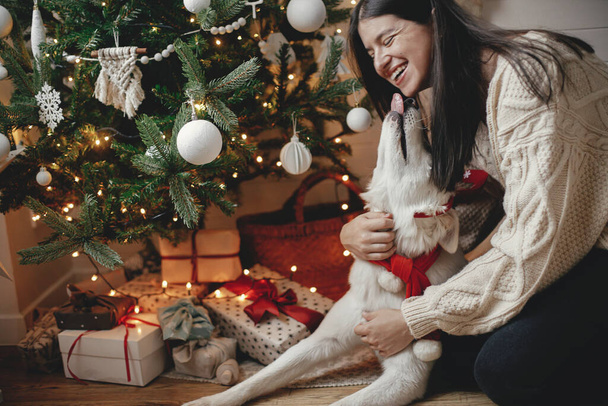 Stylish woman in cozy sweater caressing adorable dog under christmas tree with gifts and lights. Happy young female hugging and kissing cute white dog in festive scandinavian room. Happy Holidays - Zdjęcie, obraz