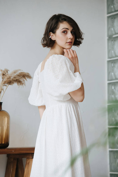 Turning back sensual shy woman in a white dress. Beautiful brunette with short hair indoors. - Valokuva, kuva