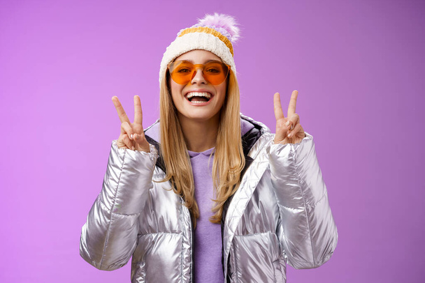 Доброзичлива оптимістична красива блондинка в сонцезахисних окулярах срібна стильна куртка зимовий капелюх показує жест перемоги у мирі, насолоджуючись засніженими канікулами, виходять за межі готелю насолоджуватися лижами, фіолетовим фоном
 - Фото, зображення
