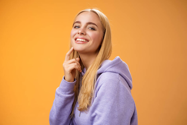 attrayant sassy auto-assuré caucasien blond femelle en violet sweat à capuche tourner caméra toucher menton cheeky sourire caméra vérifier chose intéressante regardant vers l'avant intrigué, debout fond orane - Photo, image
