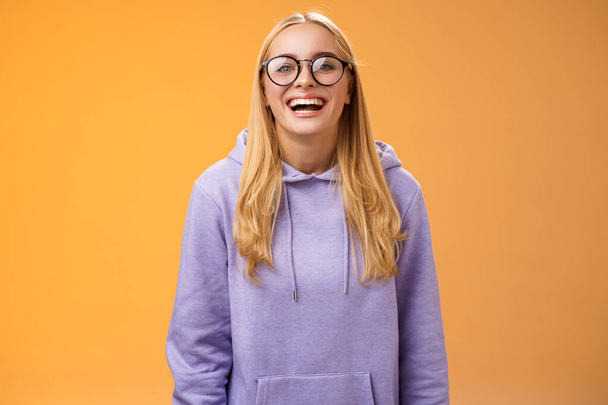 Karismaattinen iloinen viehättävä hymyilevä naispuolinen yliopisto suora opiskelija lasit violetti viihtyisä huppari hymyilee nauraen iloisesti kutsuttu käsi luokkatoverit seisoo oranssi tausta - Valokuva, kuva