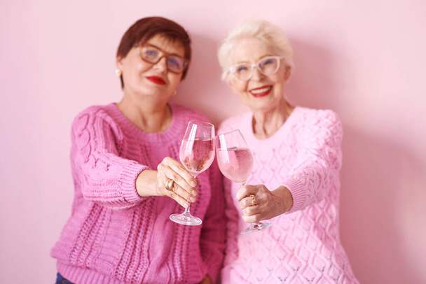 kaksi tyylikästä vanhempaa naista vaaleanpunaisissa villapaidoissa, joissa on ruusuviinilasit vaaleanpunaisella taustalla. Ystävyys, puhua, juorut, suhteet, uutiset, perheen käsite  - Valokuva, kuva