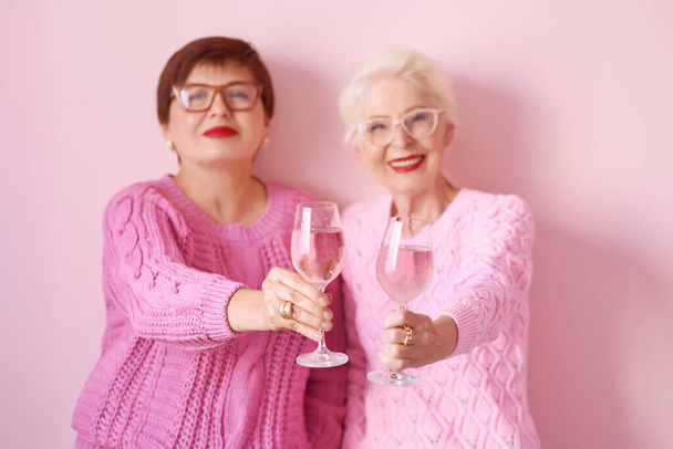 two stylish senior women in pink sweaters with rose wine glasses on pink background. Friendship, talk, gossip, relationships, news, family concept  - Photo, Image