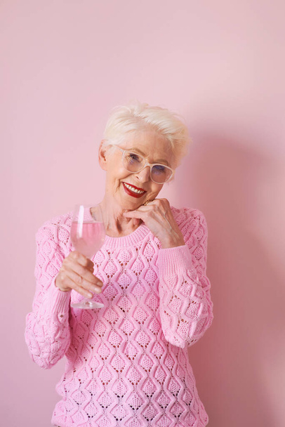 szczęśliwa starsza kobieta w kaszmirowym różowym swetrze pijąca różę na różowym tle. Świętowanie, miłość, emerytura, dojrzała koncepcja  - Zdjęcie, obraz