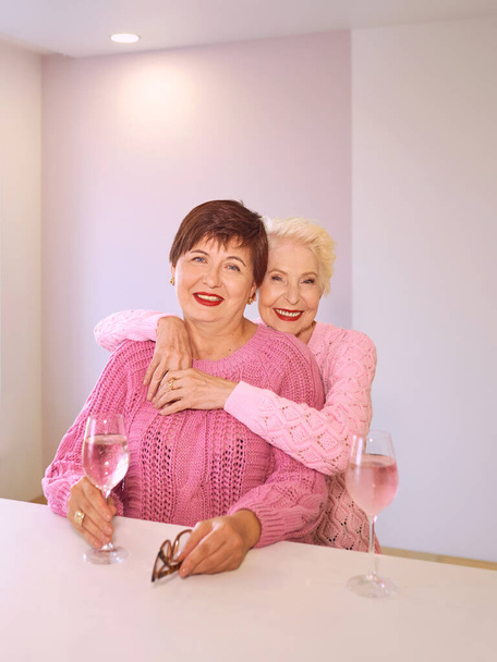 two stylish senior women in pink sweaters sitting with wine glasses in bar talking. Friendship, talk, gossip, relationships, news, family concept  - Foto, immagini