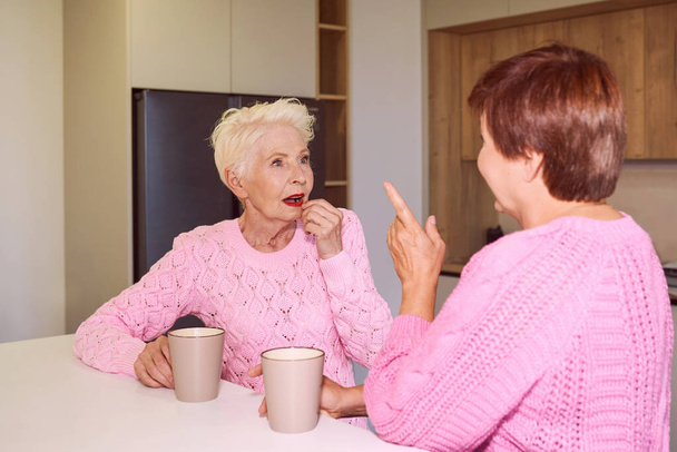 dvě stylové starší ženy v růžových svetrech sedící s hrnky u moderních kuchyňských drbů. Přátelství, řeči, drby, vztahy, novinky, rodinný koncept  - Fotografie, Obrázek