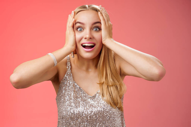 Amused surprised excited young blond attractive woman in silver glittering stylish dress hold head hands widen eyes speechless look camera unbelievable thrill fan see famous person, red background - Photo, Image