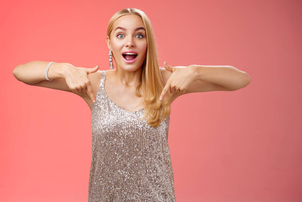 Impressed excited attractive glamour blond girl in silver glittering dress gasping thrilled pointing down glance camera fascinated check out fabulous awesome promo, standing amused red background - Photo, Image