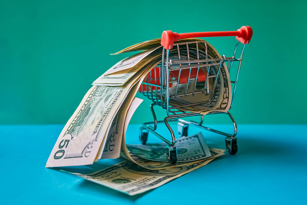 concept - curled dollar banknotes fall out of Shopping Cart Trolley against the green background - Foto, afbeelding