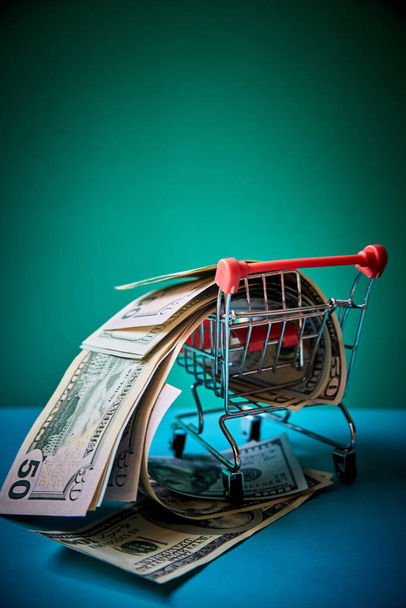 concept - curled dollar banknotes fall out of Shopping Cart Trolley against the green background - Фото, изображение
