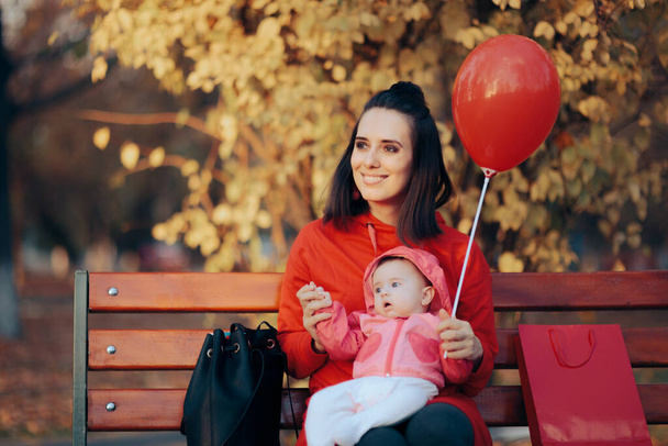 Mother Sitting on a Bench Holding Her Baby and a Red Balloon - Photo, image