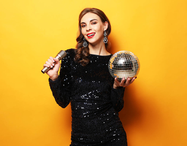Party, holiday and celebration concept: Young brunette woman with long curly hair dressed in evening dress holding a microphone and disco ball - Photo, Image