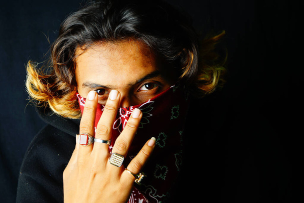 A closeup of a stylish Indian male wearing a red bandana over his face posing on the dark background - Фото, изображение