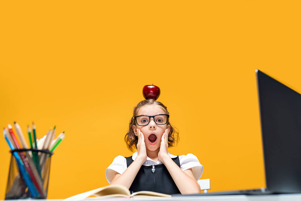 Schoolgirl in fun sitting at the desk with laptop holding apple on her head. Distance education  - 写真・画像